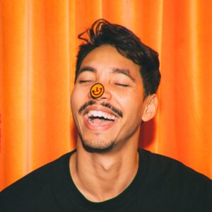 Image description: Headshot of man wearing a black tshirt against an orange background. He has a smiley face sticker on his nose.