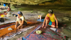 Three women on the ground outside, in a mossy, green-tinted cave, each weaving mats. They're working on very colorful mats, one orange, one blue and pink and one green. They're using rocks to hold the mats down as they weave them together.