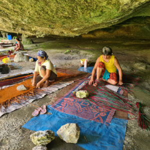 Three women on the ground outside, in a mossy, green-tinted cave, each weaving mats. They're working on very colorful mats, one orange, one blue and pink and one green. They're using rocks to hold the mats down as they weave them together.
